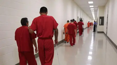 Getty Images Migrant detainees in red jumpsuits walk down a hall at an ICE detention centre in Adelanto, California in 2013 that was run by GEO Group 