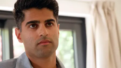 Close-up shot of Hartesh Battu looking above the camera. He has short dark hair, dark eyes and eyebrows, and slight dark stubble. He is wearing a denim shirt and is standing in front of a window, framed by thick cream curtains.