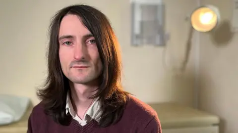 Tom sits sitting in front of an examination table and lamp. He has long, dark-brown hair and is wearing a white shirt and a burgundy V-neck jumper. 