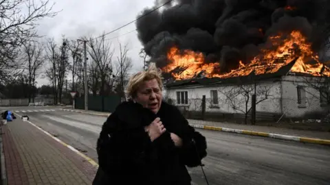 Getty Images A woman holds her black fur coat, eyes closed and mouth open, as the roof of a home behind her burns