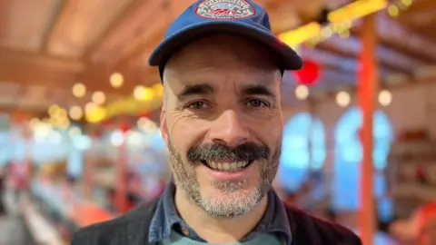 Tom Herbert. He has dark grey facial hair and is wearing a blue cap, a turquoise jumper and a shirt with a denim collar poking out. He is looking directly at the camera and smiling. 