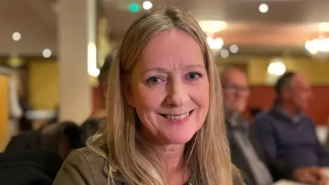 Ben Schofield/BBC Jacqueline Allen smiling and looking direct to camera. She has blonde, straight hair falling over her shoulders and blue eyes. She is wearing a dark green jacket. Other diners in a restaurant can be seen out of focus in the background. 