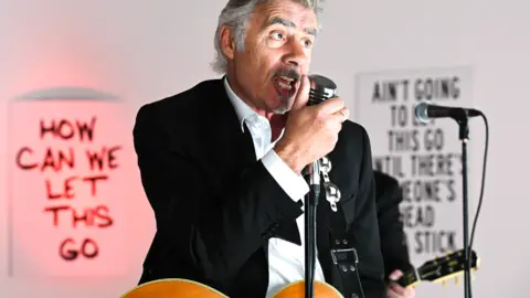 Danny Clifford Glen Matlock sings into a microphone, with a guitar around his neck, with slogans on the wall behind him, including 'How can we let this go'