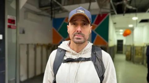 Daniel Burguet looks straight at the camera, wearing a navy baseball cap and a rucksack on his back, standing in a warehouse