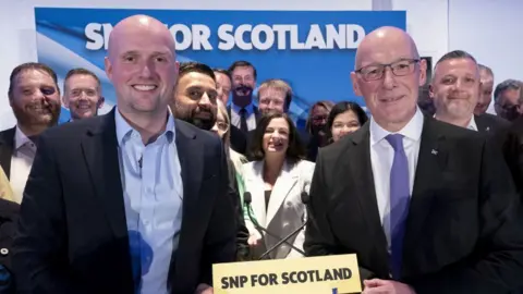 PA Media Stephen Flynn, a bald man wearing a dark suit and blue shirt, and John Swinney, a bald man wearing glasses and a dark suit with a white shirt and purple tie, smile at the camera in front of SNP colleagues at a party press conference 