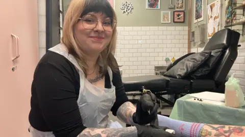 BBC Kim Thomas in her studio smiling at the camera, holding a needle and wearing black gloves and a plastic apron with a customer's tattooed legs visible in front of her 