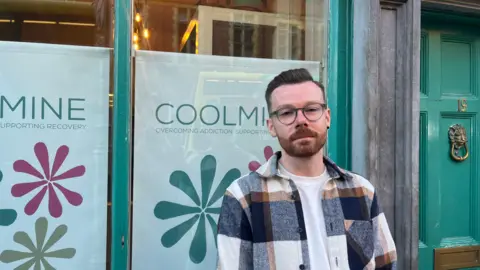 Alan Kinsella wears round grey glasses, he is wearing a shite t-shirt underneath an orange, navy and white chequered overshirt. He is standing infront of a an old green door with a gold knocker in the middle 