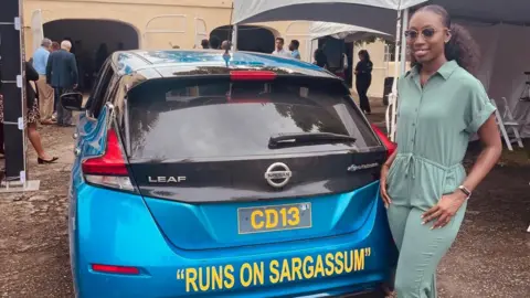 BBC Biologist Shanika Spencer poses next to a blue Nissan Leaf car on which the sentence 