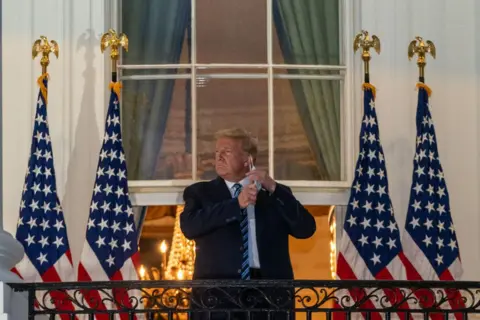 Bloomberg/Getty Images Donald Trump, in a thick, dark overcoat, removes his protective mask on the White House balcony in Washington in October 2020. Four US flags are draped behind him.