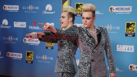 Reuters Jedward point at the camera in metallic suits in front of a blue background on the MTV Awards red carpet