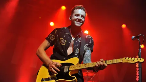 C Brandon/Redferns Danny Jones smiling while playing an electric guitar on stage. He is wearing a black flowery shirt. Red lights brighten up the background behind him