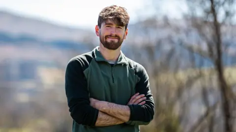 Nature Friendly Farming Network Rhys Evans, director of the Nature Friendly Farming Network in Wales, standing in a rural area, with his arms folded and smiling at the camera. He is wearing a green jumper with black on the arms. He has dark brown hair and a brown beard and moustache.