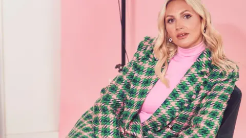 Bater and Street Sophie Pycroft looking at the camera on a chair, sitting slightly sideways. She is wearing a checked skirt suit in green and pink and a pink top.