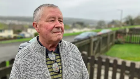 BBC Dave Cradduck stands in a garden overlooking the town of Whitehaven. He has a stern expression and he is looking at something out of shot. He has short grey hair and is wearing a grey woollen cardigan over a green and blue polo shirt. A pair of glasses are hanging from the collar of his top. 