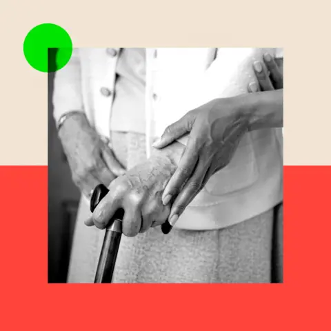 BBC A black-and-white image of an elderly woman dressed in a long skirt and blazer, holding a walking stick, as a carer gently supports her by holding her hand.