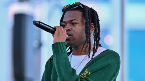 Getty Images Jordan Adetunji is standing on stage performig. He's holding a mic and is wearing a green cardigan and a pearl necklace. There are sunglasses on top of his head. 