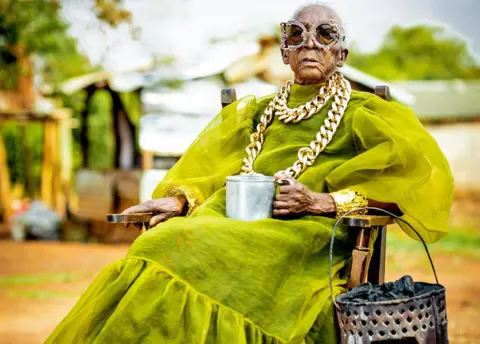 Luxury Media Zambia A seated Margret Chola wearing a chunky gold chain over a green taffeta like dress with puffy sleeves and a big frill for the hem holds a tin mug in her hand. Hanging off the arm of her chair is a 