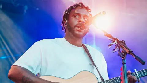 Getty Images Myles Smith performs on the Greenpeace stage during day four of Glastonbury Festival 2024 at Worthy Farmon June 29, 2024 in Glastonbury, England. He is wearing a white tshirt and has a guitar in his hands while singing into a black microphone. A spotlight behind him shines down.