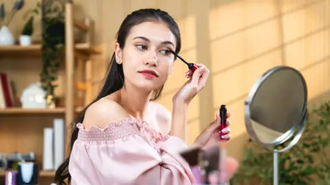 Getty Images Woman with long dark hair, wearing a pink off-the-shoulder top looking in a small mirror while she applies mascara