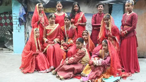 Innovation for Change A group of Indian children at a fashion show on a street, all wearing colourful red outfits and ornate jewellery including Maang Tikka, necklaces, earrings and bracelets. 