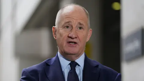 PA Media Kevin Hollinrake, who has short grey hair and wears a navy suit with a blue shirt and black tie, speaks to reporters outside a BBC building in London