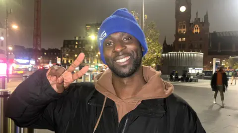 DJ AG wearing a blue beanie had doing a peace sign with his fingers. He's wearing a brown hoodie covered by a black coat with AG on the breast. There's a clocktower behind him in the main square by King's Cross with people passing by.