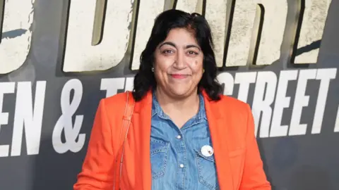 PA Media Gurinder Chadha, had long, dark hair, wearing an orange blazer and a denim shirt underneath