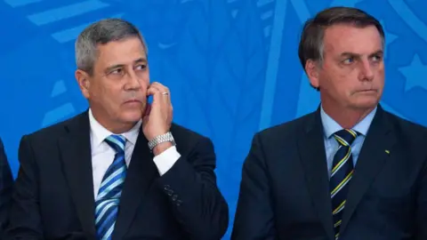 Getty Images Braga Netto, wearing a suit and tie, scratches his face as he and Jair Bolsonaro, look on during a government event in 2020