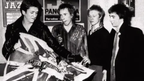 PA Media The Sex Pistols - left to right, Steve Jones, Johnny Rotten, Paul Cook and Glen Matlock - after the announcement that they have split with their record company EMI in 1977. Steve Jones, the guitarist, is tearing up a poster for their first single, Anarchy in the UK, which was released on EMI