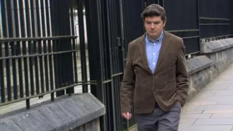 BBC Michael McMonagle walking along the pavement beside a low stone wall with tall black metal railings. He is walking with one hand in his pocket, looking down. He is wearing a brown jacket, blue shirt, and grey suit trousers. 