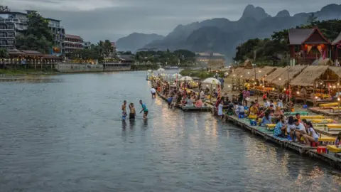 Getty Images Wide view of a river in Vang Vieng. Three people can be seen in the river while crowds are seen sitting along the river front. Mountains can be seen in the background with the town's skyline.