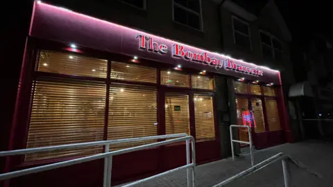 Ben Schofield/BBC An exterior shot of the Bombay Brasserie. The front of the restaurant is painted purple, with the letters of The Bombay Brasserie written in red and back-lit. There are several windows, which all have blinds over them, through which yellow light is seeping out.