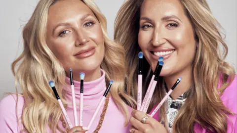 Bater and Street Sophie, left, and Hannah, right pose holding four make-up brushes each. They are wearing pink tops and the brush handles are light pink, with blue tips on the brush fibres.