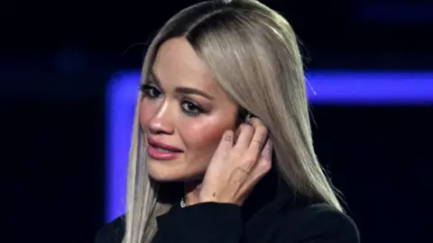 Getty Images Rita Ora in a black suit, with one hand to her ear