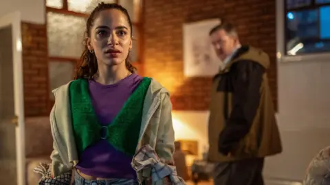 Channel 4 A young woman dressed in bright, worn clothes is staring into the distance. An man, who is of focus in the background, is staring at her. Both appear to be in a living room.