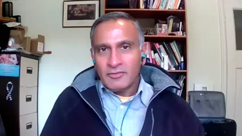 Still from an interview done on a computer recording of consultant plastic surgeon Dalvi Humzah. He has short dark greying hair swept and a tanned face and is wearing a pale blue shirt and darker blue fleece as he looks to camera.