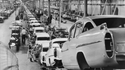 Getty Images A black and white photo of a long car production line in a large warehouse. The cars in the foreground have no wheels and workers can be seen working on the vehicles