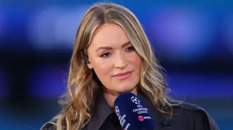 Getty Images Laura Woods holding a TNT Sports navy microphone while presenting the Champions League. She has wavy blonde hair and is wearing a black coat