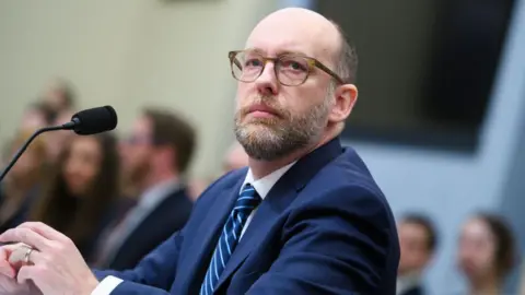 Getty Images Russell Vought is seated during a hearing with a microphone direct toward him. He is wearing a blue suit and a stipped blue tie. He is wearing glasses. 