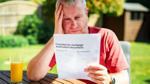 Getty Images Man looks at mortgage application documents while sitting at a garden table with a glass of orange juice in front of him. There is a lawn and shed in the background. 