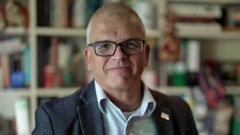 Headshot of Tim Ash, who has a slight smile, with short grey hair and black framed glasses. He is wearing a grey suit jacket and open-necked pale blue shirt. He has a small metal pin of the Ukrainian flag on the lapel of his jacket. Behind him is a wall of bookshelves. 