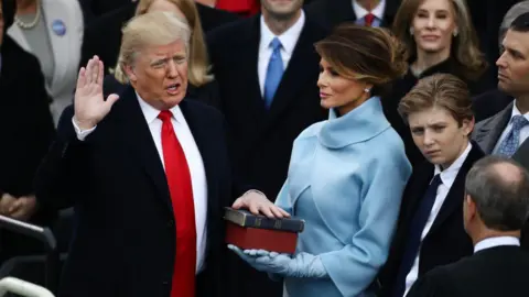 Bloomberg via Getty Images Trump wears a dark suit and red tie. He has one hand resting on a Bible and holds the other up as he recites the oath of office. Melania Trump is dressed in light blue and holds the Bible. They are surrounded by a crowd of people.