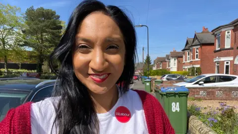 Satvir Kaur Satvir Kaur is standing in a rediential street on a sunny day, she is taking a selfie, wearing a white t shirt with a circular red Labour sticker on the left hand side and a red knitted cardigan over her shoulders.