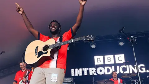 Myles Smith performing on the BBC Introducing Stage at Radio 1's Big Weekend in Luton. Myles is wearing a red Luton football jersey with a white stripe, and brown trousers. He has a guitar around his neck and both arms pointed up in the sky. Behind him are two members of the band and 