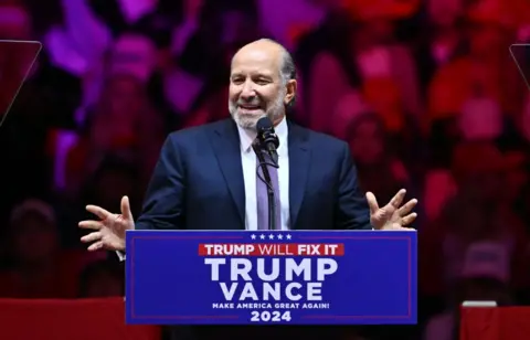 Reuters Howard Lutnick speaks while gesturing with both hands in front a lectern at at a Trump rally in Madison Square Gardens in October. He has grey hair and a beard, and wears a navy suit, white shirt and purple checkered tie.