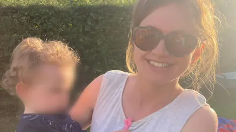 Family handout A woman holding a toddler in a garden. She has brown hair tied back and is wearing sunglasses and a grey vest top, and is smiling at the camera. The child has blonde curly hair and their face has been blurred out. There is grass and a green hedge behind them.