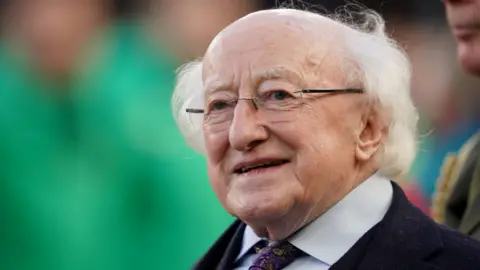 PA Media Michael D Higgins - an older man with white hair wears rimless glasses as he looks beyond the camera. He is standing with his mouth open and is wearing a dark coat, a light coloured collared shirt and a purple and yellow paisley print tie.