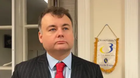 A man dressed in a black pin-stripe suit, a blue shirt and a red tie, stands in front of a doorway