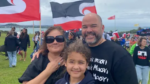 Juliet Tainui-Hernandez A family of three smiles at the camera, while people wave flags in the background