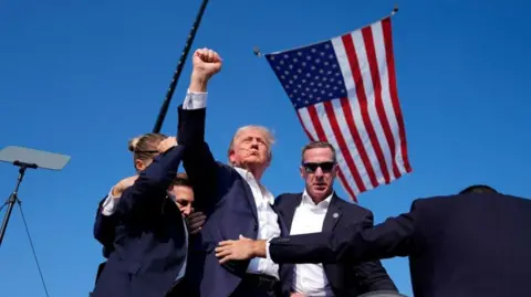 Evan Vucci/AP Donald Trump raises his fist as he is surrounded by US Secret Service agents after the shooting